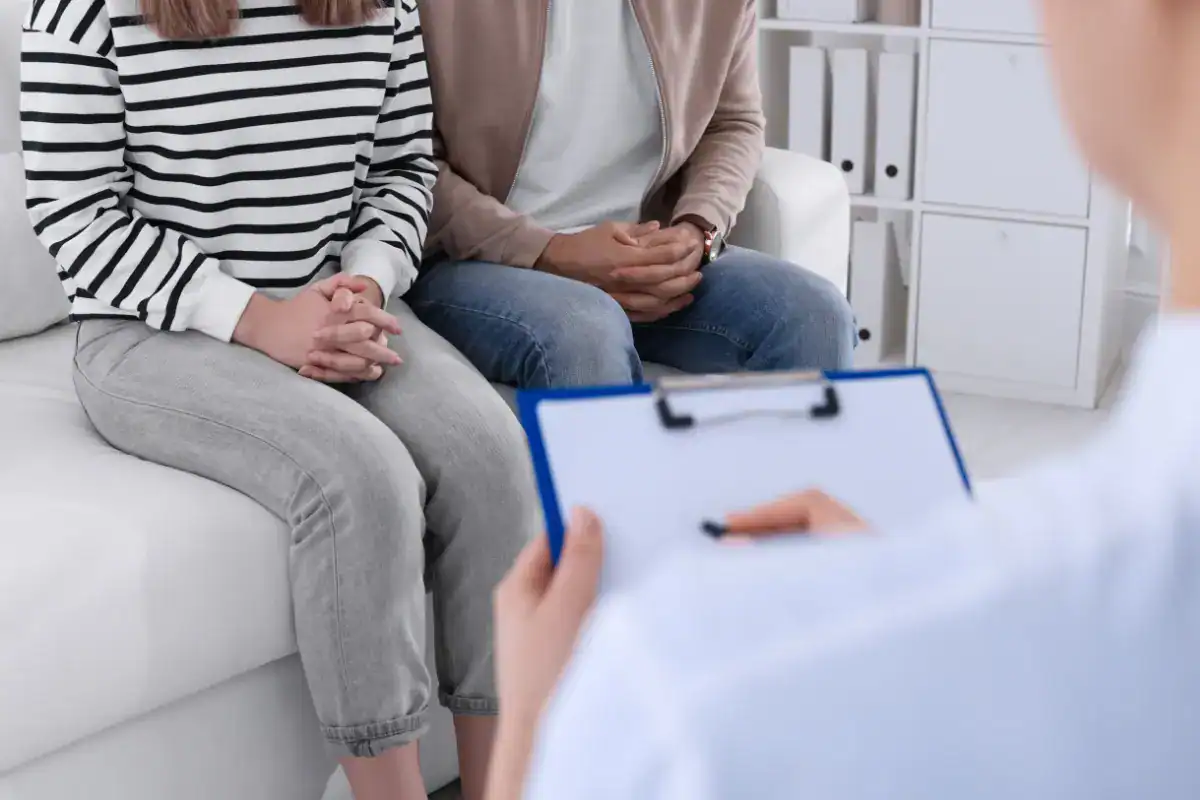 A man and woman sitting side by side on a couch with a doctor holding a clipboard out of focus in the foreground